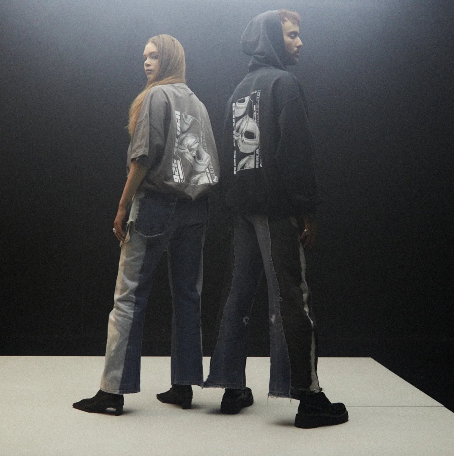 Zwei Personen in Streetwear mit auffälligen Jeans und bedruckten Oberteilen vor dunklem Hintergrund