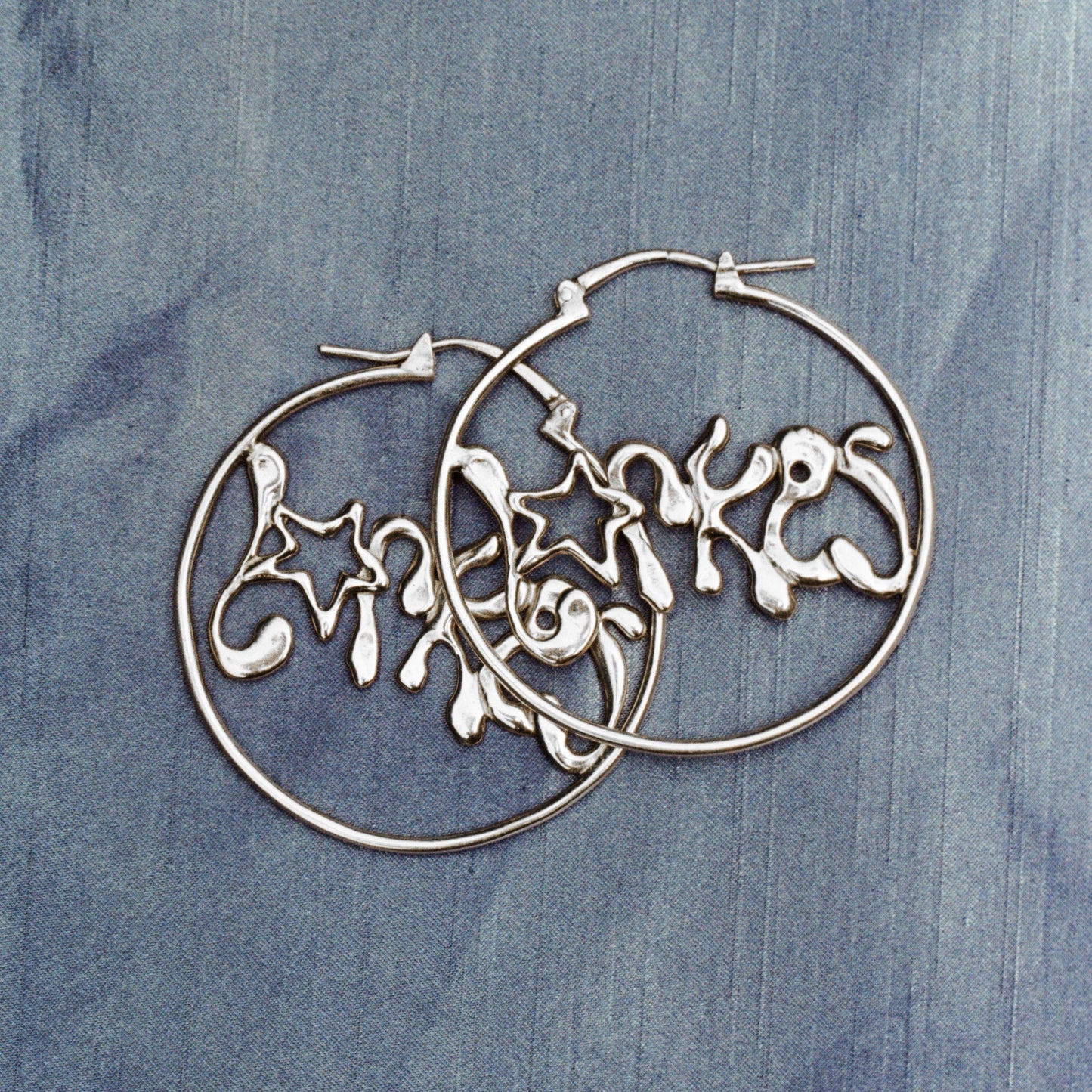 Silberne Creolen-Ohrringe mit verspieltem Stern-Design auf blauem Stoffhintergrund.