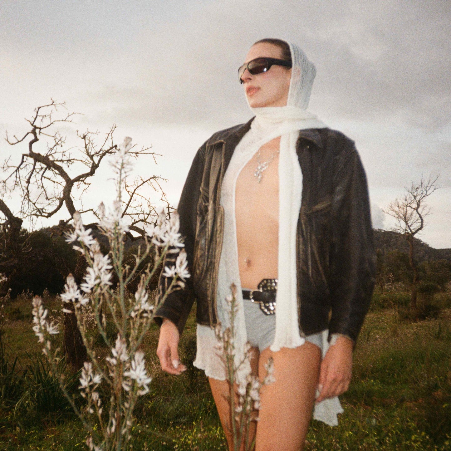 Frau mit silbernem Pferdeanhänger, Lederjacke und Sonnenbrille in natürlicher Landschaft