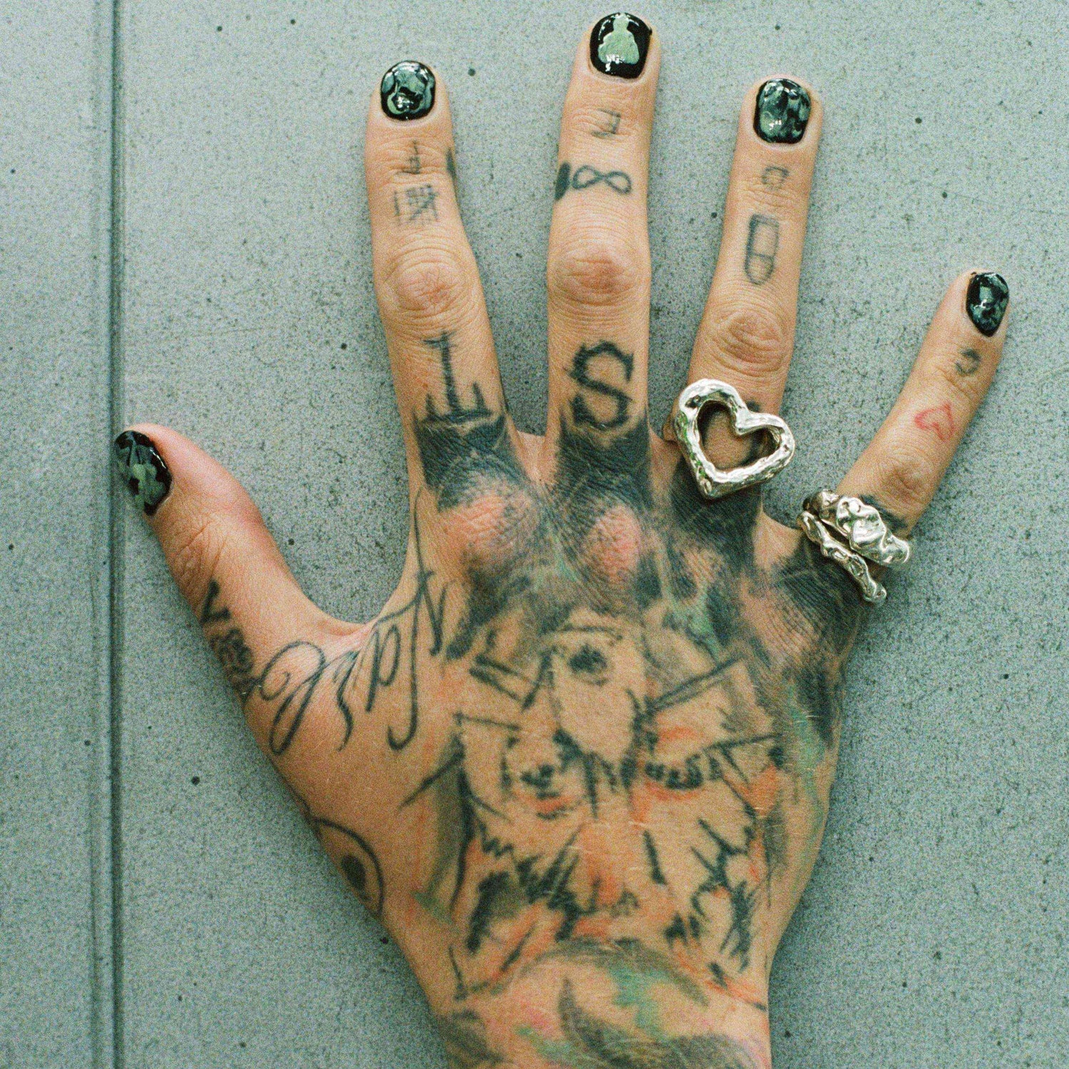 Hand mit Tattoos und silbernen Ringen, darunter ein auffälliger Herzring, vor grauem Hintergrund